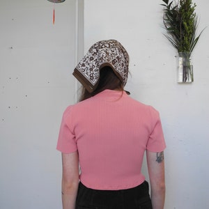 May include: A pink ribbed short-sleeve top paired with dark corduroy pants. A brown and white patterned scarf covers the head. A decorative lantern hangs in the background, along with a vase of greenery.