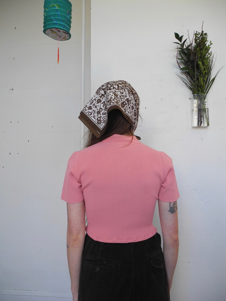 May include: A pink ribbed short-sleeve top paired with dark corduroy pants. A brown and white patterned scarf covers the head. A decorative lantern hangs in the background, along with a vase of greenery.
