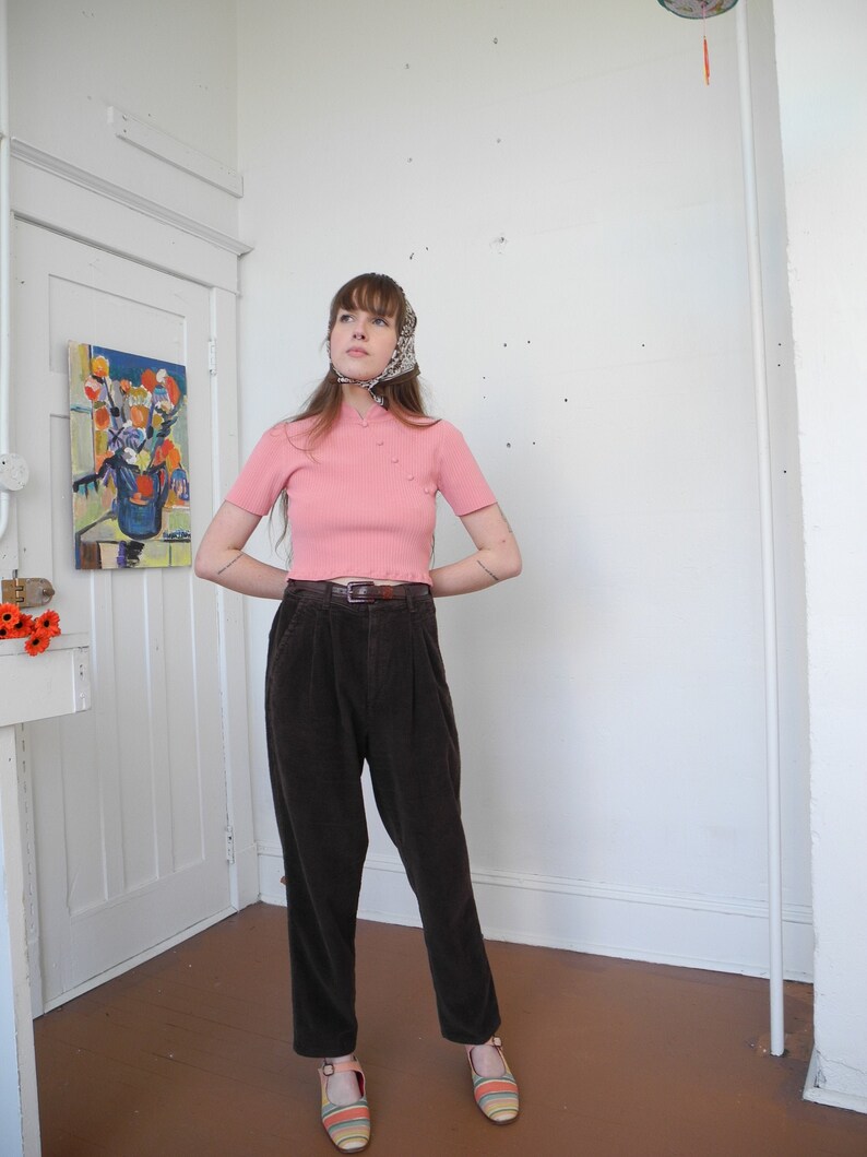 May include: A woman wearing a light pink short-sleeved top with small pearl-like buttons, paired with dark brown corduroy pants and striped sandals. A patterned scarf is tied around the head. The outfit is completed with a brown belt.