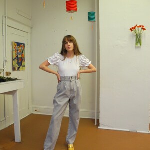 May include: A woman wearing a white puff-sleeve shirt and striped high-waisted pants. She is standing in a room with white walls and a wooden floor. There are three paper lanterns hanging from the ceiling.