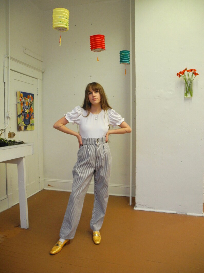 May include: A woman wearing a white puff-sleeve shirt and striped high-waisted pants. She is standing in a room with white walls and a wooden floor. There are three paper lanterns hanging from the ceiling.