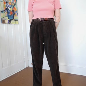 May include: A light pink ribbed knit top with a mandarin collar and three buttons. The top is paired with dark brown corduroy pants and a brown belt. The outfit is completed with striped flats.