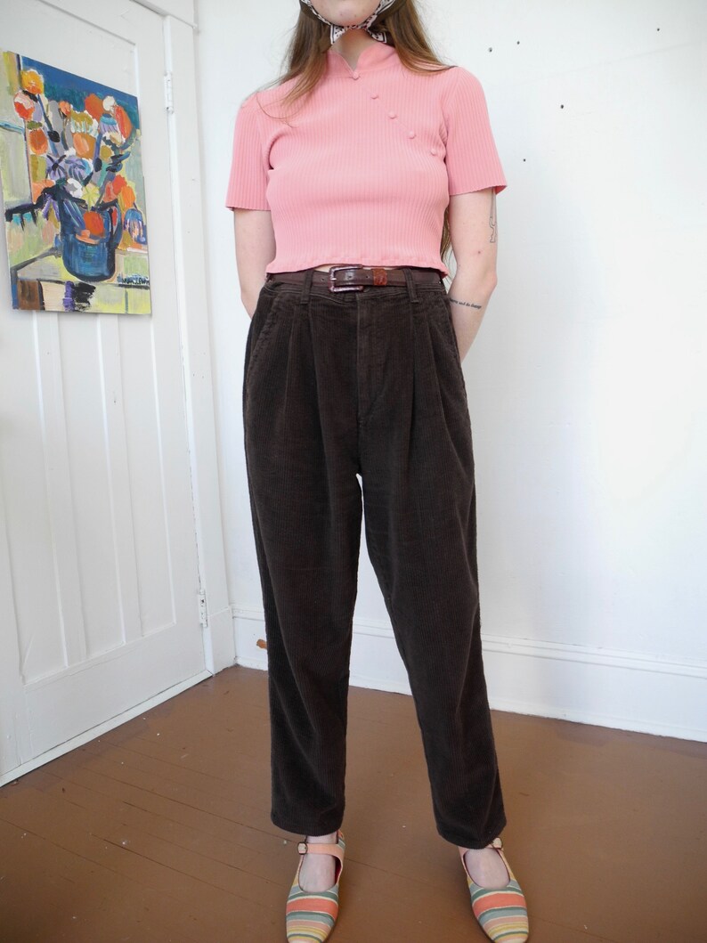 May include: A light pink ribbed knit top with a mandarin collar and three buttons. The top is paired with dark brown corduroy pants and a brown belt. The outfit is completed with striped flats.