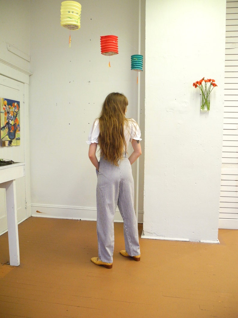 May include: A person wearing a white blouse and blue and white striped pants. The person is standing in a room with a white wall and a wooden floor. There are three paper lanterns hanging from the ceiling.