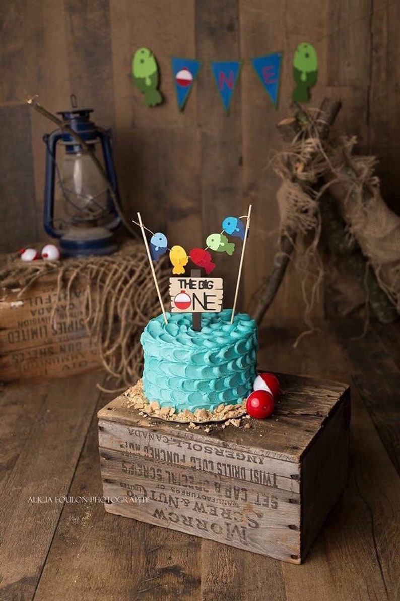 The Big One Cake Topper  unique first birthday party ideas - shown: fishing themed cake with blue wave icing and bobbers with a string of fish pennant.