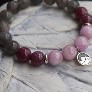 May include: A beaded bracelet featuring a mix of round, polished gemstones in shades of gray, burgundy, and pink. The bracelet includes a silver charm with a mountain design. The beads are strung together, creating a stylish accessory.