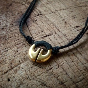 Pode incluir: Um pingente dourado com um design único pendurado em um cordão preto. O pingente é um círculo com uma divisão no meio, criando duas asas.