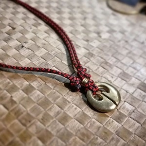 Puede incluir: Un collar de cuerda rojo y negro con un colgante de latón con un diseño estilizado. El colgante es redondo y tiene una hendidura en el medio.