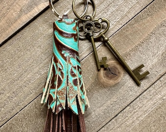 Op de afbeelding: Een handgemaakte sleutelhanger met een kwast van turquoise en bruin leer, versierd met een decoratief patroon. De sleutelhanger bevat twee sleutels in antieke stijl en een houten accent, bevestigd aan een zilveren ring.
