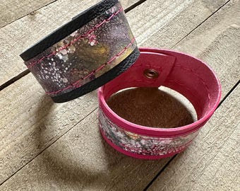 Può includere: Due polsini in pelle. Uno è nero con un design astratto colorato e cuciture rosa. L'altro è rosa acceso con un design simile e una chiusura a scatto. I polsini sono su una superficie di legno.