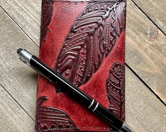 Könnte beinhalten: Ein rotes Lederjournal mit geprägten Federdesigns und einem schwarzen Stift. Das Journal hat eine detaillierte, strukturierte Oberfläche und ist geschlossen. Der Stift ist schwarz mit silbernen Akzenten.