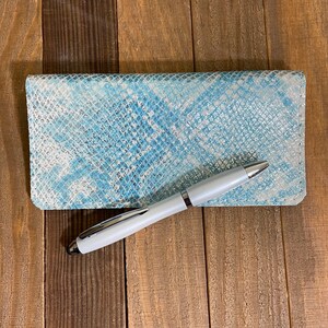 May include: A light blue and white snakeskin-patterned checkbook cover with a white pen. The cover is rectangular and has a textured surface. The pen is white with silver accents.