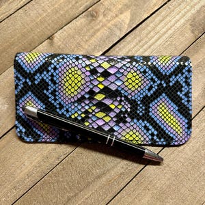 May include: A rectangular wallet with a snakeskin pattern in shades of blue, purple, yellow, and black. A black pen with silver accents rests on top of the wallet, which is placed on a wooden surface.