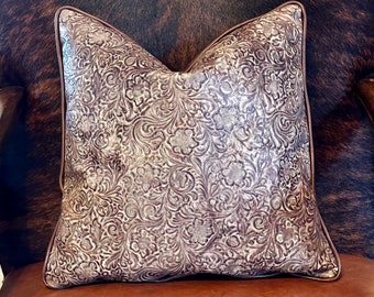 May include: A brown leather pillow with a white embossed floral pattern. The pillow is sitting on a brown leather chair.