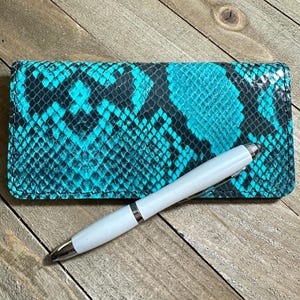 Peut inclure: Une couverture de chéquier en peau de serpent turquoise et noire avec un stylo argenté sur le dessus.