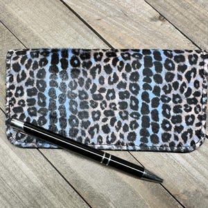 May include: A rectangular wallet featuring a textured animal print pattern in shades of black, white, and light blue. A sleek black ballpoint pen with silver-toned accents and a stylus tip is positioned diagonally across the front of the wallet.
