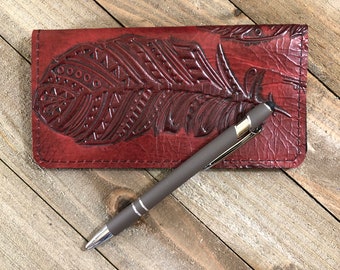 Puede incluir: Una cartera de cuero burdeos con un diseño de pluma cincelada. Un bolígrafo plateado está encima de la cartera.