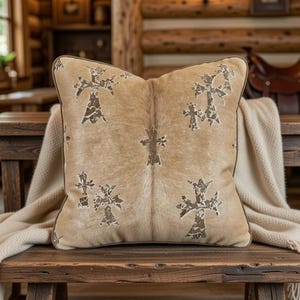 Puede incluir: Un cojín decorativo cuadrado con un patrón de piel de vaca en tonos beige y marrón. El cojín presenta un diseño de cruz repetido en un color marrón más oscuro. El cojín está colocado sobre un banco de madera con una manta color crema.