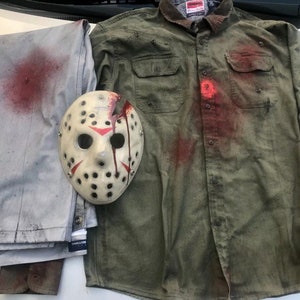 May include: A white hockey mask with red paint splatters and a green button-up shirt with red paint splatters. The shirt has a pocket on the left side and a button-up collar. A pair of grey pants with red paint splatters are folded on the left side of the image.