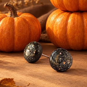 May include: A pair of round stud earrings featuring a glossy black surface with embedded gold-colored flecks. The earrings are positioned in front of three orange pumpkins and scattered dried autumn leaves on a brown surface.