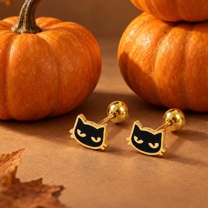May include: A pair of gold-tone stud earrings featuring a black cat face design with a ball backing, positioned near three orange pumpkins and dried autumn leaves. The cat faces have gold outlines and simple, stylized facial features.