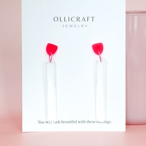 Könnte beinhalten: Ein Paar durchsichtige Acryl-Ohrringe mit rosa Oberteilen. Die Ohrringe werden auf einer weißen Karte mit dem Text "OLLICRAFT JEWELRY" und "Sie werden mit diesen Ohrringen wunderschön aussehen" präsentiert.