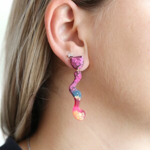 Puede incluir: Un par de pendientes de color rosa, azul y dorado con purpurina y un dise&ntilde;o ondulado abstracto.