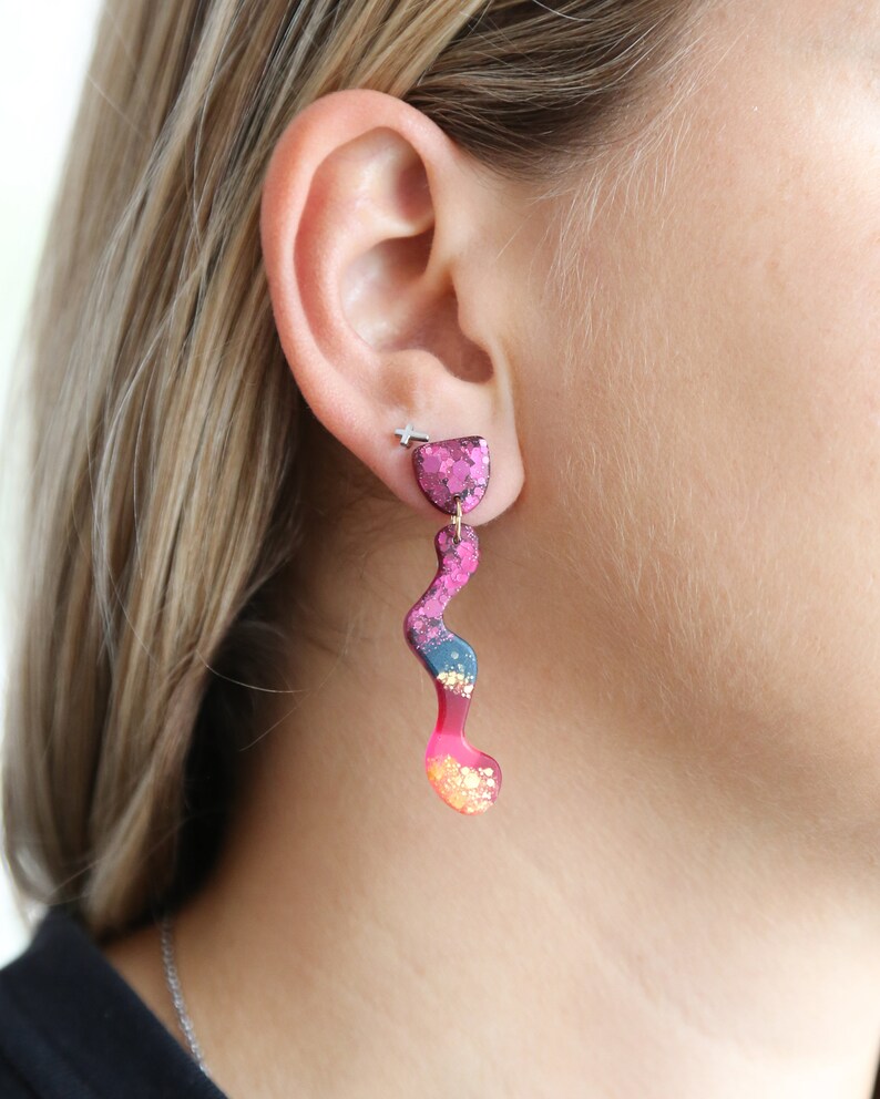 Puede incluir: Un par de pendientes de color rosa, azul y dorado con purpurina y un dise&ntilde;o ondulado abstracto.