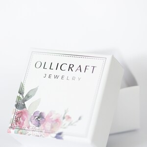 May include: White jewelry box with the text "OLLICRAFT JEWELRY" in silver lettering. The box features a floral design with pink, purple, and green watercolor flowers. The box is slightly open, revealing the inside.