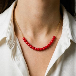 May include: A delicate necklace featuring a strand of small, vibrant red beads. The beads are strung on a fine gold chain, creating a simple yet elegant design. The necklace is shown against a white shirt.