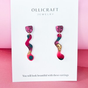 Puede incluir: Un par de pendientes colgantes abstractos de color rosa, negro y dorado con un acabado brillante. Los pendientes est&aacute;n sobre una tarjeta blanca con el texto "OLLICRAFT JEWELRY" y "Te ver&aacute;s hermosa con estos pendientes".