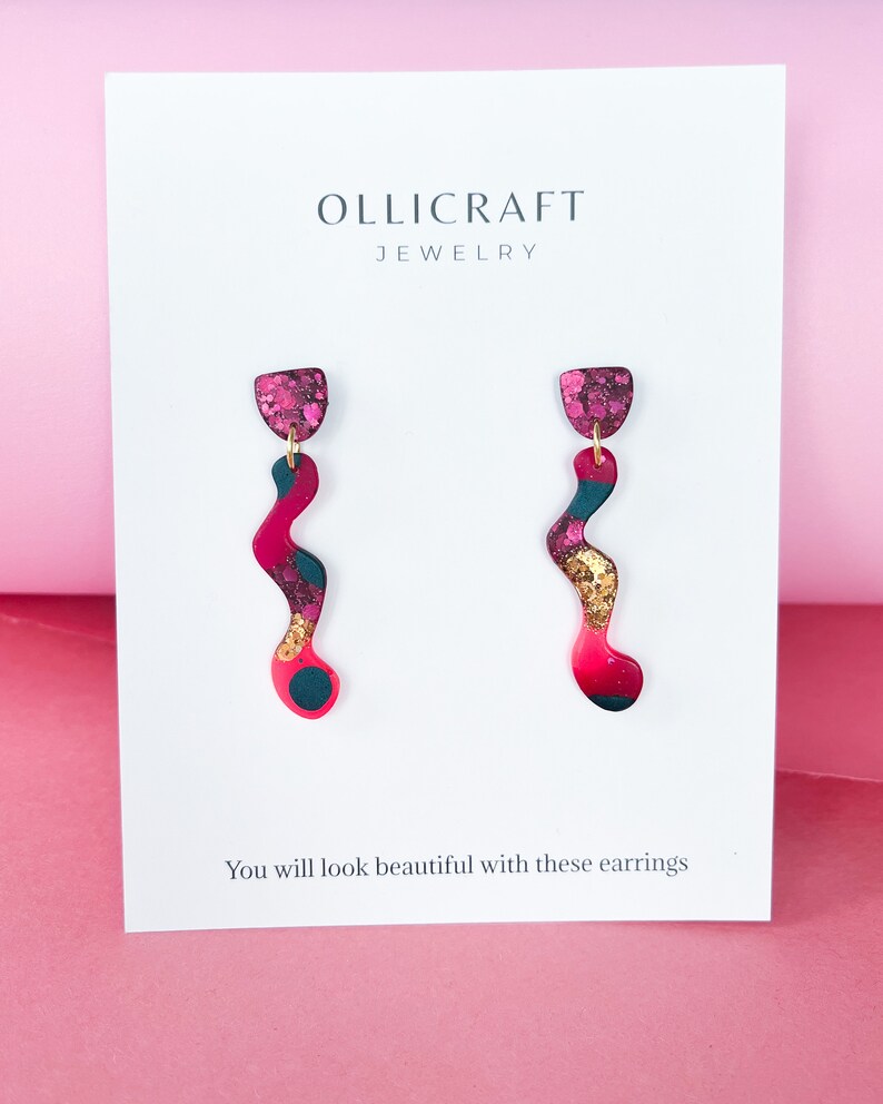 Puede incluir: Un par de pendientes colgantes abstractos de color rosa, negro y dorado con un acabado brillante. Los pendientes est&aacute;n sobre una tarjeta blanca con el texto "OLLICRAFT JEWELRY" y "Te ver&aacute;s hermosa con estos pendientes".