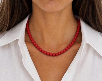 Puede incluir: Un collar compuesto por una sola hilera de pequeñas cuentas redondas de color rojo mate. Las cuentas son uniformes en tamaño y forma, ensartadas para formar una cadena circular continua que descansa en la base del cuello, sobre una camisa blanca.