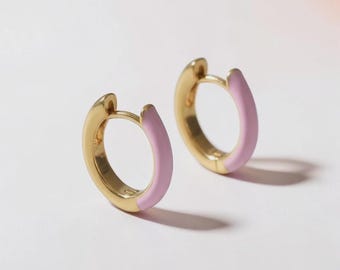May include: A pair of small gold hoop earrings featuring a smooth, light pink enamel coating on the outer half of each circular band. The earrings have a hinged closure mechanism and a polished metallic finish on the inner side of the hoops.