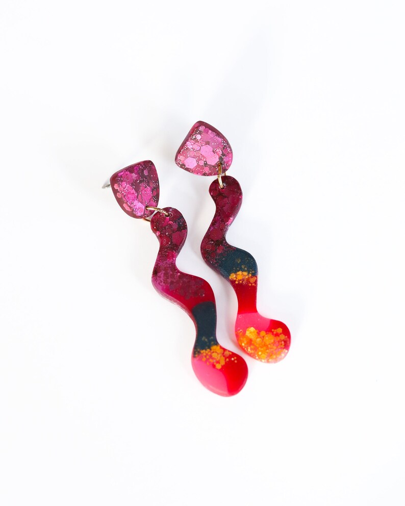 Puede incluir: Un par de pendientes largos, abstractos, de color rosa, negro y dorado con un dise&ntilde;o ondulado. Los pendientes est&aacute;n hechos de resina y tienen un acabado moteado.