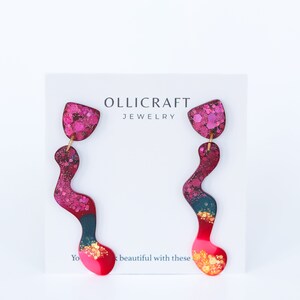 Puede incluir: Un par de pendientes largos, abstractos, de color rosa, rojo y azul con detalles de purpurina dorada. Los pendientes est&aacute;n sobre una tarjeta blanca con el texto "OLLICRAFT JEWELRY".