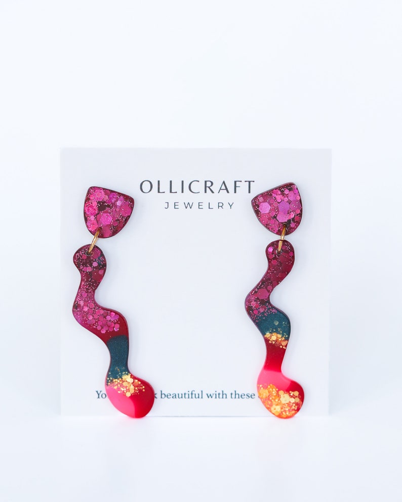 Puede incluir: Un par de pendientes largos, abstractos, de color rosa, rojo y azul con detalles de purpurina dorada. Los pendientes est&aacute;n sobre una tarjeta blanca con el texto "OLLICRAFT JEWELRY".