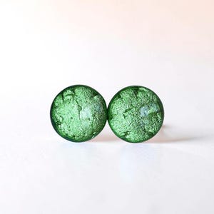 May include: Two round, textured, green glass stud earrings. The earrings have a slightly irregular surface, catching the light. The earrings are set against a plain white background. These earrings are a simple, elegant accessory.