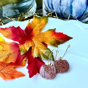 May include: A pair of gold hoop earrings with a sparkly pumpkin charm. The earrings are displayed on a white surface with fall leaves.