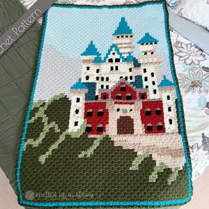 Peut inclure: Une couverture au crochet représentant un château coloré avec un toit bleu et des accents rouges. Le château est entouré d'arbres verts et d'un ciel bleu. La couverture est bordée d'une garniture turquoise. "Knitted in a Stitch" est écrit dans le coin inférieur.