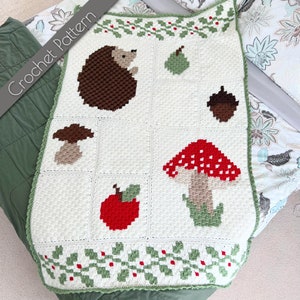 Puede incluir: Una manta de crochet blanca con un borde verde y un patrón repetido de hojas de acebo y bayas. La manta presenta una variedad de imágenes de crochet, incluyendo un erizo, un hongo, una manzana y una pera.