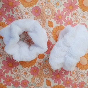 Puede incluir: Dos scrunchies de pelo blancos y peludos sobre un fondo floral.