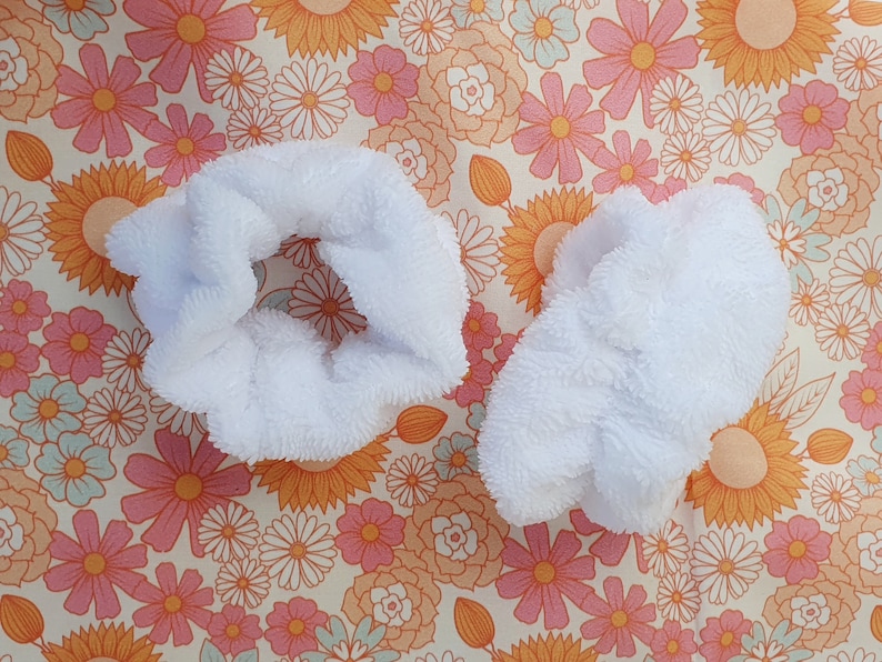 Puede incluir: Dos scrunchies de pelo blancos y peludos sobre un fondo floral.