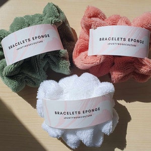 Puede incluir: Tres scrunchies suaves y esponjosos en verde, rosa y blanco. Cada scrunchie tiene una etiqueta blanca con el texto "BRACELETS EPONGE #DUSTYROSECOUTURE" impreso en ella.