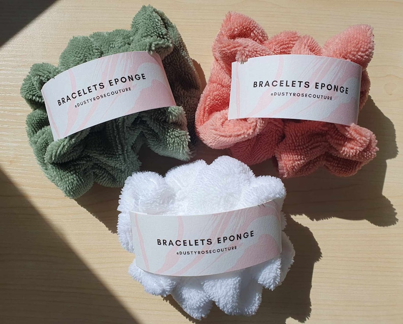 Puede incluir: Tres scrunchies suaves y esponjosos en verde, rosa y blanco. Cada scrunchie tiene una etiqueta blanca con el texto "BRACELETS EPONGE #DUSTYROSECOUTURE" impreso en ella.