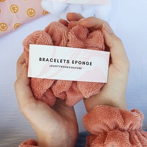 Puede incluir: Dos scrunchies de color rosa esponjoso en las mu&ntilde;ecas de una persona. Los scrunchies est&aacute;n etiquetados con "Bracelets Eponge" y "@dustyrosecouture".