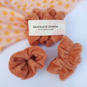 Puede incluir: Tres scrunchies marrones hechos de tela suave y esponjosa. Los scrunchies est&aacute;n dispuestos sobre una superficie blanca. Un tejido rosa con caritas sonrientes amarillas est&aacute; en el fondo. Una etiqueta blanca con el texto "BRACELETS EPONGE #DUSTYROSECOUTURE" es visible en la imagen.