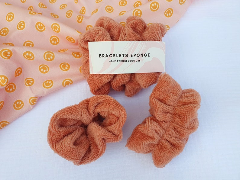 Puede incluir: Tres scrunchies marrones hechos de tela suave y esponjosa. Los scrunchies est&aacute;n dispuestos sobre una superficie blanca. Un tejido rosa con caritas sonrientes amarillas est&aacute; en el fondo. Una etiqueta blanca con el texto "BRACELETS EPONGE #DUSTYROSECOUTURE" es visible en la imagen.