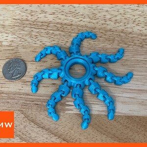 May include: A bright blue, 3D printed, articulated, star-shaped object with a central ring. The object is made of multiple connected segments, resembling a chain or a series of links. It is similar in size to a US quarter.