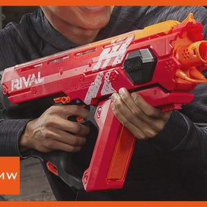 May include: Red and orange toy blaster with the word "RIVAL" printed on the side. The blaster has a black grip and the text "MXV-5000" is visible. The blaster is held by a person wearing a gray long-sleeve shirt.